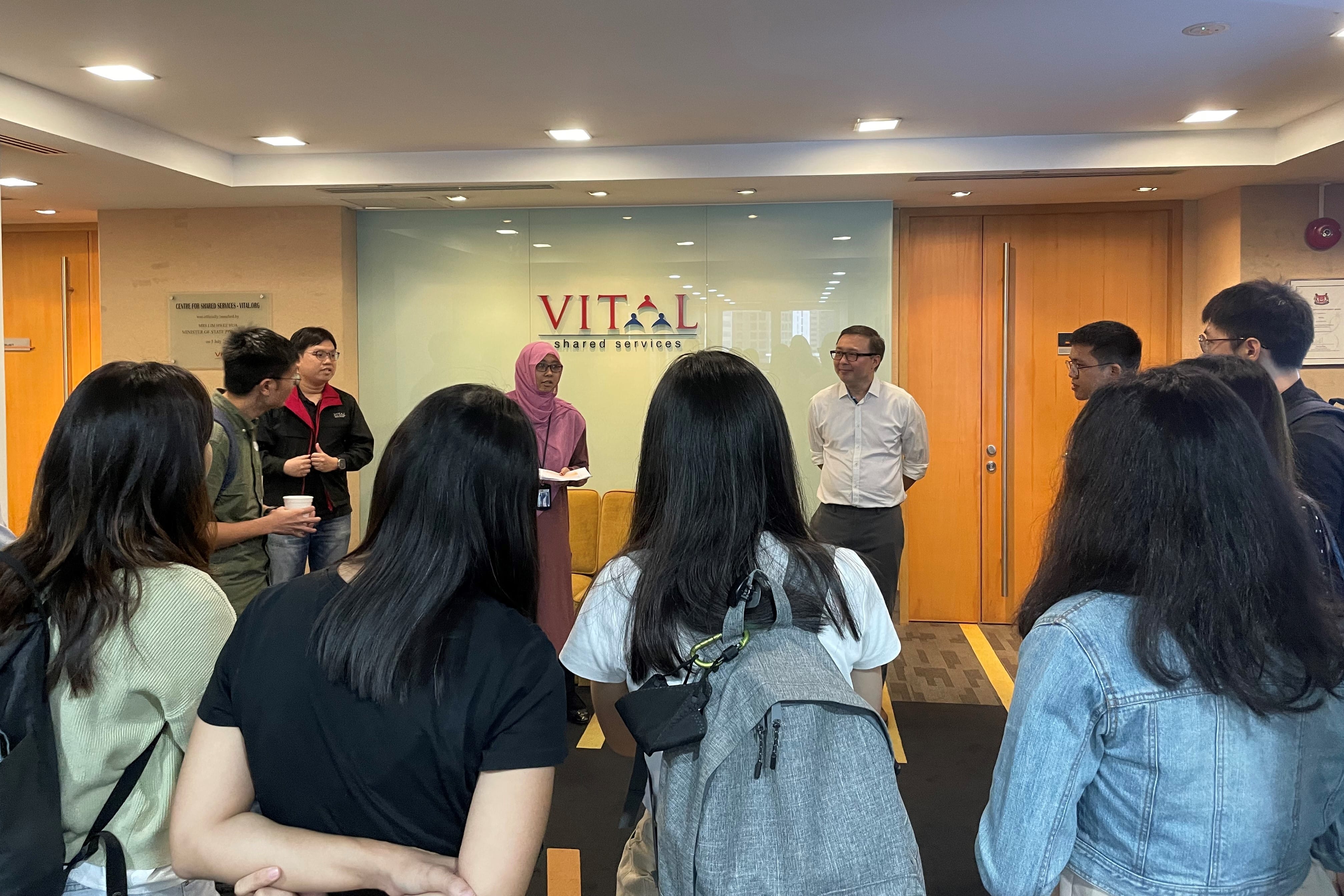 A group of people gather in the lobby of Vital Shared Services office, listening to two speakers near the entrance.